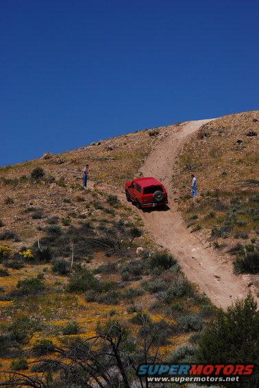 Red SUV with spare tire on rear door, driving up a dirt hill with two people standing nearby
