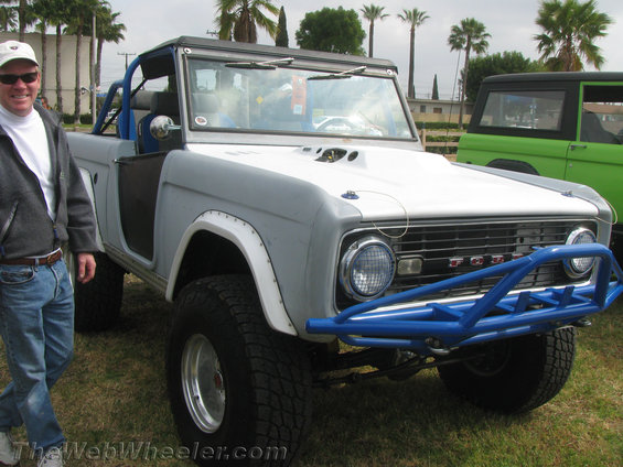 Gray Ford Bronco with blue roll cage and custom bumper, parked next to a green Bronco
