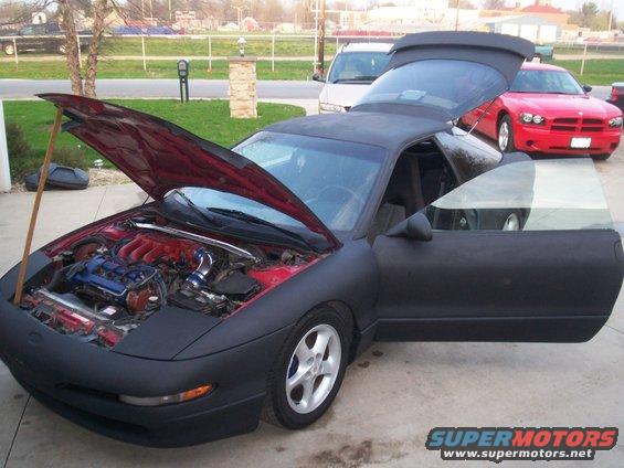 Black 1993 Ford Probe with hood and driver door open, showing engine. Red Dodge Charger in background