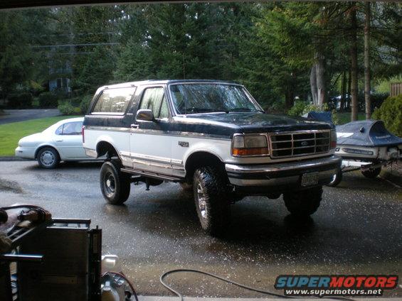 lifted two-tone Ford Bronco parked on wet pavement, sedan and jet ski trailer nearby