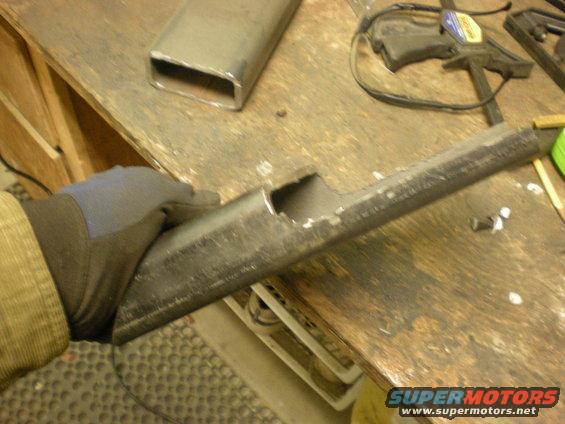 Gloved hand holding a metal tube with a rectangular cut-out, resting on a wooden workbench