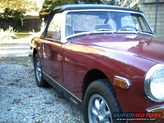 Maroon convertible sports car parked on gravel. Has a black soft top