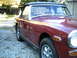 1973 MG Midget - This maroon convertible sports car is parked on a gravel driveway with trees in the background.