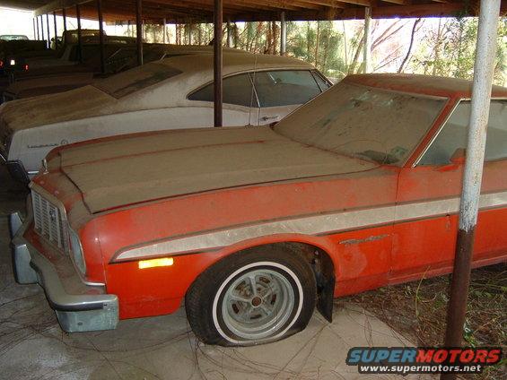 Orange 1977 Ford Ranchero with a flat tire, covered in dust, parked under a shelter