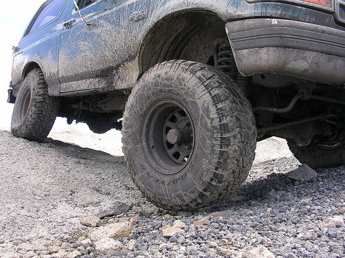 Mud-splattered dark green 1996 Ford Bronco side view, showing large off-road tires and suspension on gravel