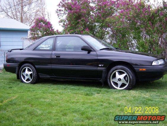 Black 1989 Mazda MX-6 GT coupe parked on grass, side view. Aftermarket wheels and a spoiler are visible