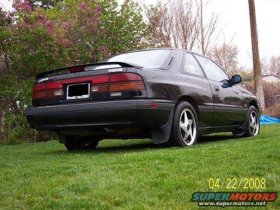 Black 1989 Mazda MX-6 coupe parked on grass with a spoiler and aftermarket wheels