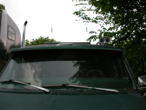 Green van front view with dual air horns on roof and a large truck to the left