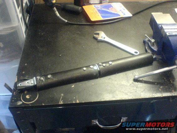 Black cylindrical part with bolts and metal bracket on workbench next to adjustable wrench and vise