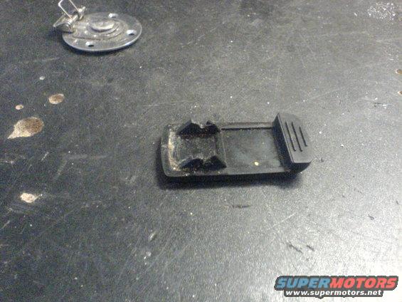 Black plastic car part with clips on a dark surface. A metal circular piece with a latch is nearby
