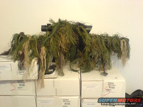 Sniper ghillie suit draped over boxes, with a scope visible on top