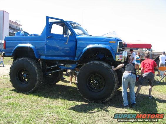 Blue lifted 1978 Ford Bronco with oversized tires and open driver's door, people examining the front wheel