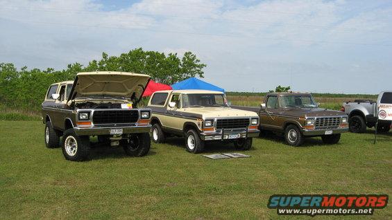 Three Ford Broncos and a pickup truck parked on grass with tents in background. One Bronco has hood open