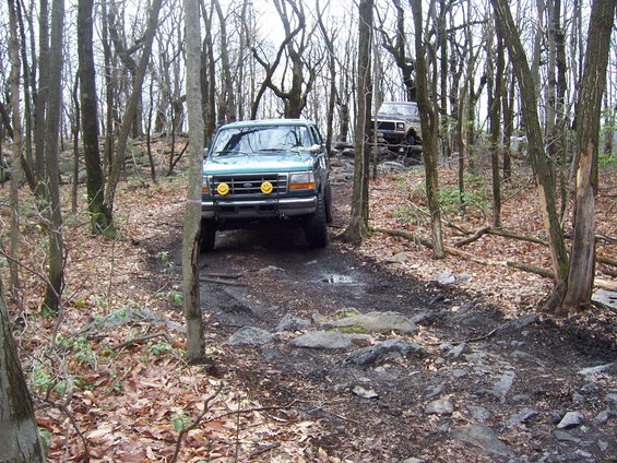Green Ford F-Series truck with smiley face lights on the bumper, driving on a rocky, wooded trail. Another SUV is behind it