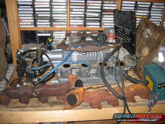 Engine block and exhaust manifolds on a wooden surface, with shelves of cassette tapes in the background