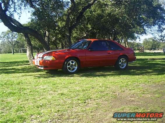 Red 1990 Ford Mustang coupe with white racing stripes on the hood, parked on grass under trees