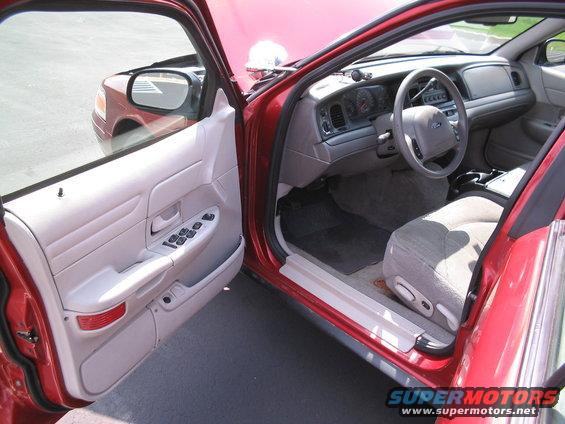 Interior of a red sedan, driver's side door open. Dashboard, steering wheel, and seats visible. Another red car is reflected in the side mirror