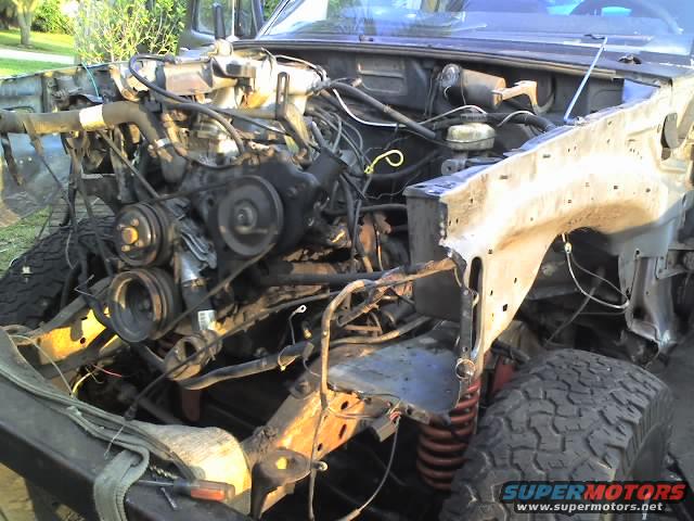 Disassembled vehicle engine bay with exposed engine, wiring, and suspension components. Off-road tires visible