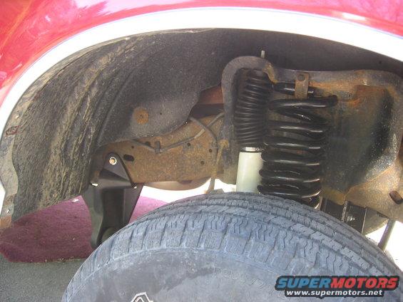 Close-up of a vehicle's front wheel well showing the tire, shock absorber, and coil spring. Text overlay reads SUPERMOTORS www.supermotors.net