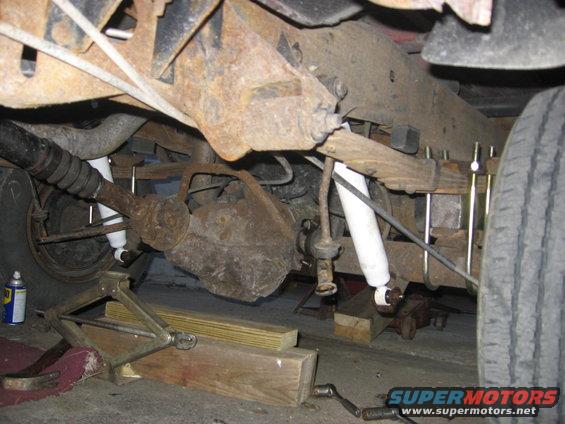 Underneath view of rusty rear axle, leaf springs, and shocks. A jack and wooden blocks are visible