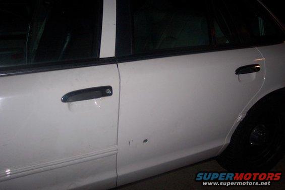Side view of a white sedan's doors and rear wheel. There are two door handles and a small dent on the lower door panel