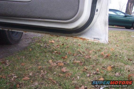 Open car door showing rusted sill. Green car visible in background