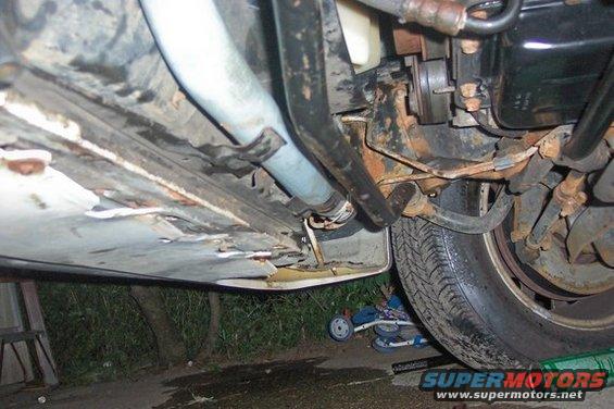 Underside of a car with significant rust and damage to the undercarriage paneling, showing a tire and suspension components