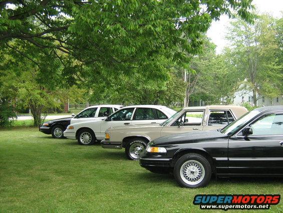 Four sedans parked in a row on a grassy lawn under trees. The closest car is black