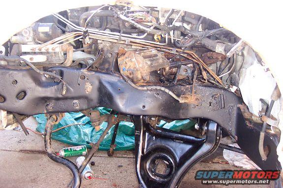 Vehicle frame with engine parts, hoses, and wiring. Some rust is visible