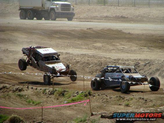 Two off-road buggies race on a dirt track; one is airborne. A large truck is in the background