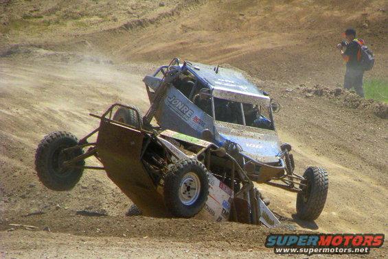 Two off-road buggies are tangled up on a dirt track, one on top of the other. A person with a backpack stands on a hill in the background