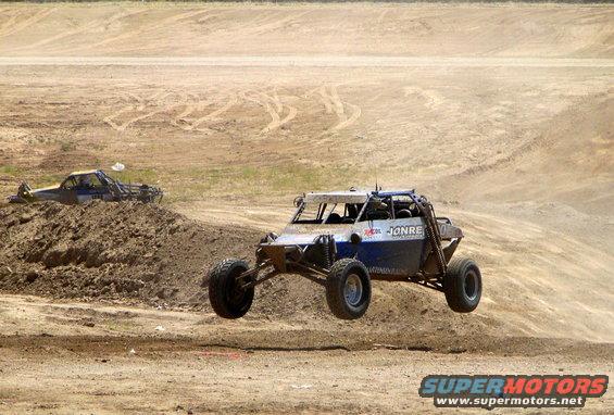 Off-road buggy mid-air jump, another vehicle stuck in dirt behind it. Text on buggy reads JONRE
