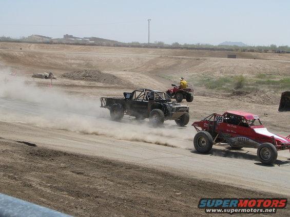 Two off-road buggies and a truck race on a dirt track, kicking up dust. An ATV follows behind