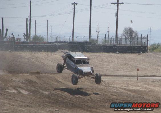 Off-road buggy mid-air over dirt track, blue and silver body, roll cage visible
