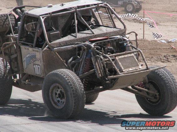 Mud-covered buggy with number 107 on the side, racing on a dirt track. Another buggy is visible in the background