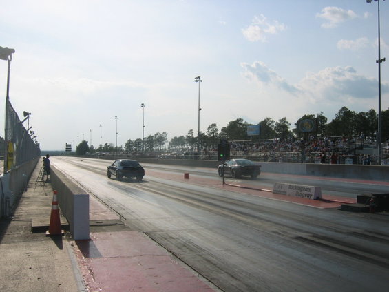 Two cars racing on a drag strip with spectators in the background. Rockingham Dragway sign visible
