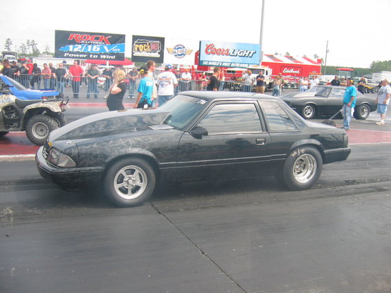Black drag car with custom hood flames on track, another car and ATV nearby