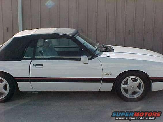 White convertible sports car parked outside, black top up. Side view shows red stripe along the bottom