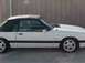 1989 Ford Mustang - A white convertible sports car with a black top is shown from the side, parked in front of a building.
