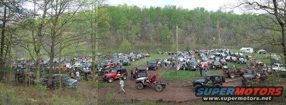 Many off-road vehicles parked in a muddy field with trees and hills in the background