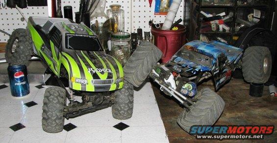Two RC monster trucks, one green and black, the other blue and black, sit on a checkered floor next to a Pepsi can