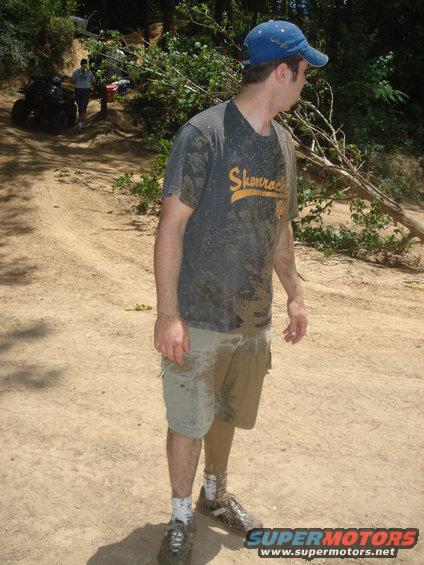 Man covered in mud standing on a dirt path with ATVs in the background