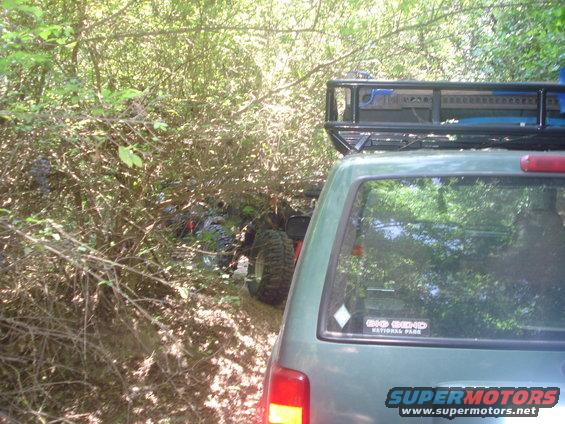 Green SUV with roof rack on a trail, another off-road vehicle visible in the trees