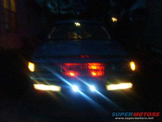 Car front at night, headlights and turn signals illuminated. Red taillights visible