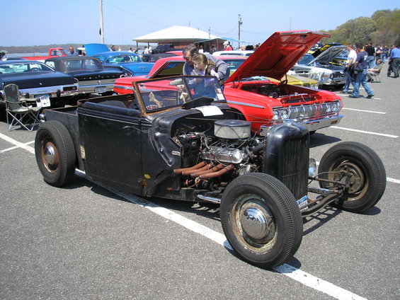 Black hot rod with exposed engine and front wheels visible. Other cars and people are in the background
