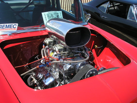 Close-up of a chrome supercharger and engine components in a red car's engine bay. Another car is visible in the background