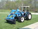 1984 Honda ATC 250 R - A blue tractor with a front loader is parked on a grassy lawn with trees in the background.