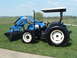 1984 Honda ATC 250 R - A blue New Holland tractor with a front loader is parked on a grassy field under a bright blue sky.