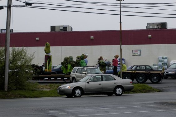 People in green costumes stand around a flatbed truck with six wheels, a statue, and other vehicles in a parking lot