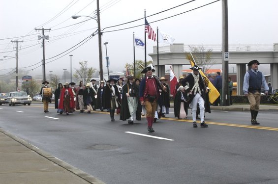Colonial reenactors march in a parade with flags and a drum